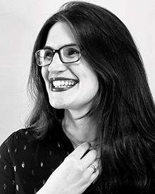This black-and-white photograph features a dark-haired woman with glasses and dark lipstick in side profile. She is smiling, and touching with her right hand the necklace she is wearing.